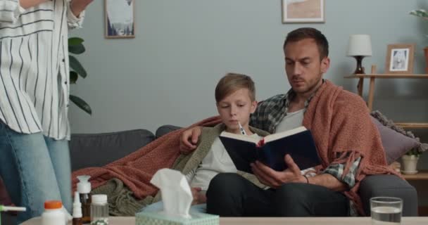 Nastolatek mierzący temperaturę i czytający książkę z tatą, gdy siedzą na kanapie. Uśmiechnięta mama biorąca i patrząca na termometr przynosząc gorący napój. Pojęcie choroby. — Wideo stockowe