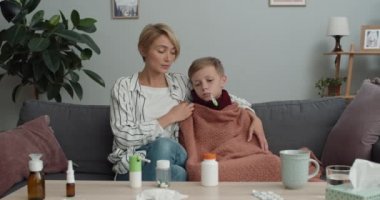Anne termometreyi tutuyor ve hasta çocukla kanepede oturup kameraya bakarken konuşuyor. Kadın ve oğlu evde doktorla çevrimiçi tıbbi konsültasyon yapıyorlar..