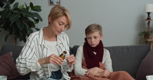 Junge Frau gießt Hustensaft ein und gibt ihrem kranken Teenager-Sohn. Kranker Junge mit Schal am Hals, der Medikamente nimmt, während er mit seiner Mutter auf dem Sofa sitzt. Konzept von Krankheit und Medizin. — Stockvideo