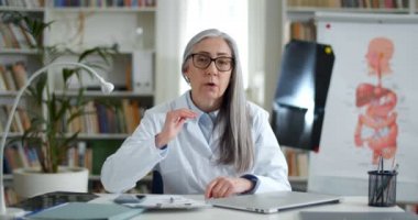Çevrimiçi danışmanlık yaparken konuşan, el kol hareketi yapan gözlüklü ve beyaz profesyonel ceketli neşeli yaşlı kadın. Kadın gülümseyen doktor sağlık odasında kameraya bakıyor..