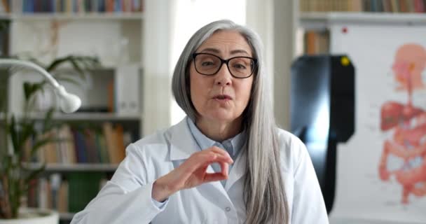 Crop view of female doctor in glasses showing ok sign and waving bye while having video consultation. Woman in white gown talking and looking to camera in medical office. Telemedicine. — Stock Video