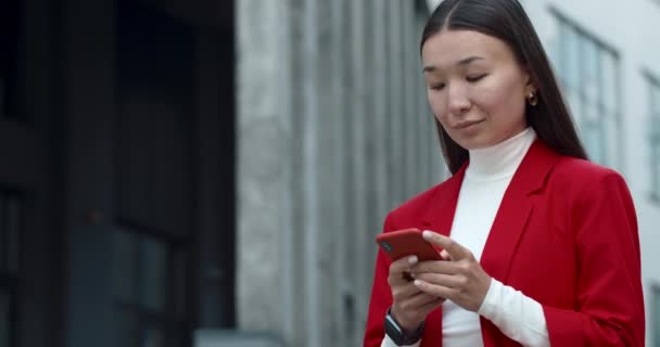 街の通りに立っている間、スマートフォンを使用して魅力的なビジネス女性。スクロールしながら携帯電話の画面に触れながら脇を見てスタイリッシュなアジアの女性。成功者の概念. — ストック動画
