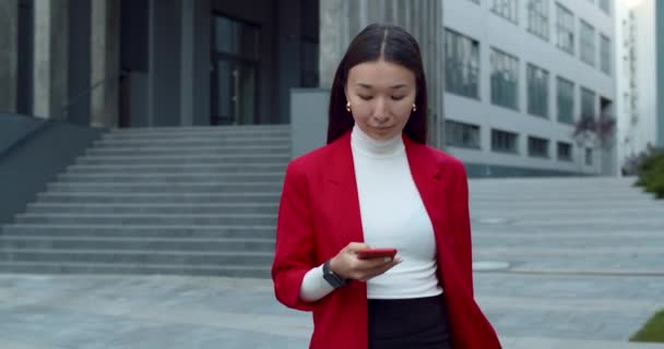 Vista de la cosecha de buen aspecto mujer de negocios asiática utilizando el teléfono inteligente y mirando a un lado. Mujer positiva exitosa tocando y presionando la pantalla del teléfono mientras camina en la calle en el área de negocios. — Vídeos de Stock