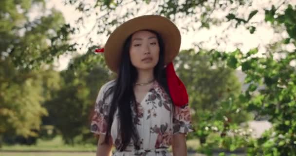 Ritratto di giovane donna asiatica che gira la testa e guarda la macchina fotografica alla luce del giorno. Bellissimo modello femminile con cappello alla moda in posa e sorridente mentre in piedi nel parco. Zoom in avanti. Tempo ventoso. — Video Stock