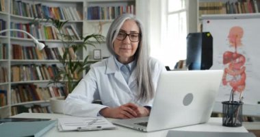 Kameraya bakıp gülümseyen olgun bir kadının portresi. Tıp ofisinde dizüstü bilgisayarın önünde otururken. Gözlüklü kadın doktor ve beyaz profesyonel elbise..
