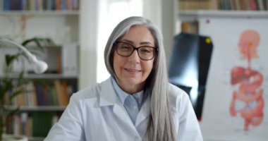 Gözlüklü ve beyaz profesyonel elbiseli bir kadının online danışmanlık yaparken el sallaması ve konuşması. Ofiste kameraya bakan kadın doktor. TeleHealth kavramı.