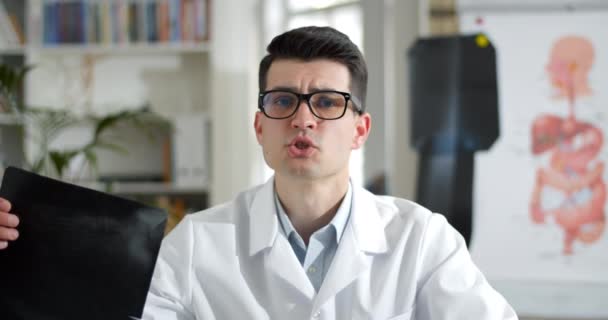 Crop view of young man in glasses and white gown showing x ray picture to patient and talking while having video call. Male doctor looking to camera in medical office. — Stock Video