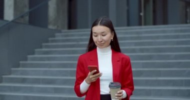 Akıllı telefon kullanan ve ofis binasının merdivenlerinden inerken kağıt kahve fincanı taşıyan pozitif bir iş kadını. Asyalı kadın gülümsüyor, yan tarafa bakıyor ve kameraya bakıyor..