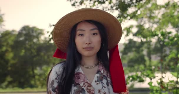 Retrato de una mujer asiática con un elegante sombrero mirando a la cámara a la luz del día. Vista de la cosecha de la mujer joven y bonita posando y sonriendo mientras está de pie en el parque. Concepto de estilo de vida. — Vídeo de stock