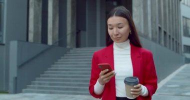 Güzel bir kadının akıllı telefon kullanması ve şehir caddesinde yürürken kağıt kahve fincanı taşıması. Asyalı bir iş kadını telefon ekranına dokunurken yana bakıyor..