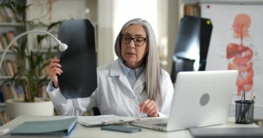 Kadın doktor, internetteki tıbbi muayenede göğüs röntgeni çekiyor. Gözlüklü ve beyaz elbiseli olgun bir kadın ofisinde hastasıyla iletişim kurarken dizüstü bilgisayar kullanıyor..