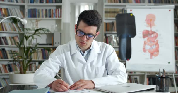 Primer plano del hombre en blanco rob llenar el formulario médico, mientras que tener video consulta en línea. Joven médico varón escuchando al paciente y escribiendo información mientras mira a la cámara. — Vídeo de stock