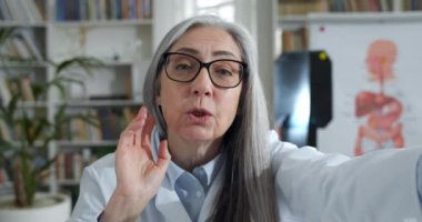 Gözlüklü, akıllı telefon tutarken el sallayan ve konuşan neşeli kadın doktorları kapatın. Ofiste online danışmanlık yaparken ekranda baskı yapan ve gülümseyen yaşlı kadın..