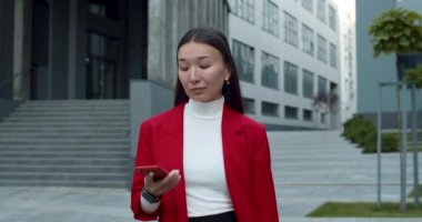 Şık giyinmiş çekici Asyalı kadın telefona cevap veriyor ve gülümsüyor. Başarılı genç iş kadını akıllı telefonla konuşuyor ve iş alanında sokakta yürüyor..