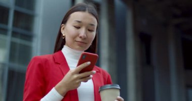 Akıllı telefon kullanan ve elinde kağıt kahve fincanı tutan bir kadının şehir caddesinde duruşu. İnternette gezinirken Asyalı iş kadını ekranı karıştırıyor ve dokunuyor.