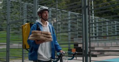 Spor sahasının yanında yürürken bisikletini iten yiyecek çantalı bir erkek kurye. Sipariş verirken pizza kutuları taşıyan koruyucu bisiklet kasklı bir adam..