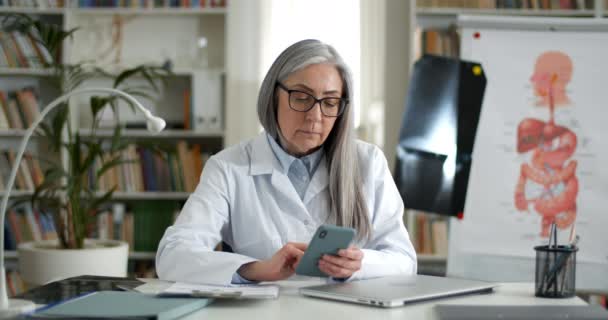 Femme âgée en lunettes et robe blanche défilement et toucher smartphone sreen. Femme médecin mature assis à table dans le bureau médical. Concept de médecine et de santé. — Video