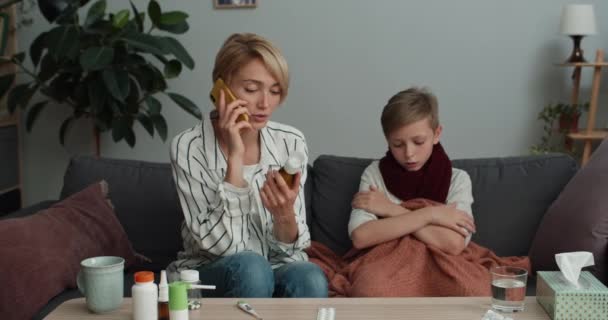 Νεαρή μητέρα κρατώντας σιρόπι για το βήχα και μιλώντας στο smartphone, ενώ κάθεται στον καναπέ με άρρωστο παιδί. Όμορφη γυναίκα επικοινωνεί με τον γιατρό στο τηλέφωνο. Έννοια της ασθένειας και της ιατρικής. — Αρχείο Βίντεο