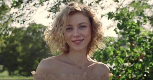 Portrait de femme heureuse regardant vers la caméra et souriant tout en soufflant le vent ses cheveux blonds. Vue rapprochée d'une jolie femme posant au soleil dans le parc. Concept de style de vie. — Video