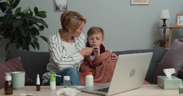 Genç bir kadın konuşuyor ve ergen çocuğuna termometre veriyor. Video sohbeti sırasında hapları gösteriyor. Anne ve oğlu, doktorla çevrimiçi danışmanlık yaparken bilgisayar ekranına bakıyorlar.. — Stok video
