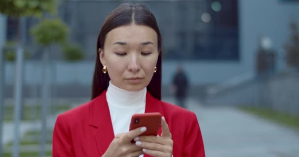 スマホを使いながらも良いニュースと笑顔を持つ魅力的なアジア人女性の姿を間近で見ることができます。成功したビジネスマンは、通りに立っている間に電話画面を見て触れます. — ストック動画