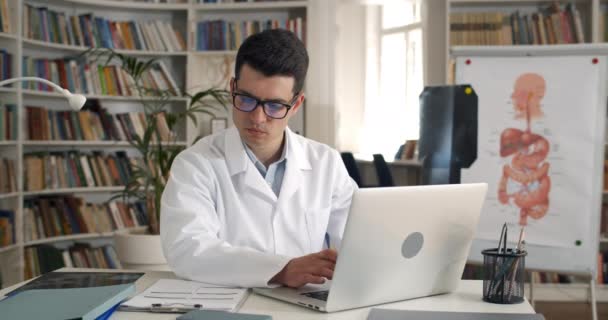 Porträt eines männlichen Arztes, der im Büro Informationen von der Krankenakte bis zum Laptop eingibt. Schnittansicht des männlichen Arztes in Brille und weißem Berufskleid am Arbeitsplatz. — Stockvideo