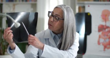 Yetişkin bir kadının gözlüklü ve beyaz önlüklü görüntüsü hastaya röntgen filmi gösteriyor ve video görüşmesi yaparken konuşuyor. Kadın doktor muayenehanede kameraya bakıyor..