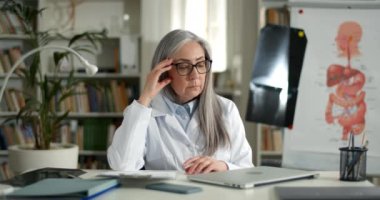 Olgun bir kadın doktor masada dizüstü bilgisayarla oturup düşünürken yan yana bakar. Gözlüklü ve beyaz Rob 'lu bir kadın. Sağlık ofisinde çalışırken düşünceli görünüyor..