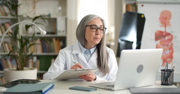 Vrouwelijke volwassen arts in medische rob met online video overleg terwijl zitten aan tafel in het kantoor. Oudere glimlachende dame praten terwijl het vasthouden van papieren houder en met behulp van laptop. — Stockvideo