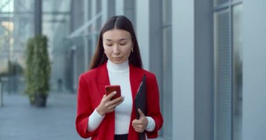 İş kadını telefon içeriğini kontrol ediyor ve şehir caddesinde yürürken mutlu görünüyor. Belge klasörünü taşırken akıllı telefon ve ekrana dokunan Asyalı çekici kadın .