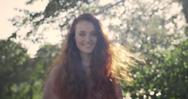 긴 머리를 하고 웃으면서 대낮에 카메라를 보는 아름다운 여성의 모습. 공원에서 서 있는 젊은 여자가 포즈를 취하는 모습이 눈에 띈다. 확대 해. 생활 양식에 대한 개념. — 비디오