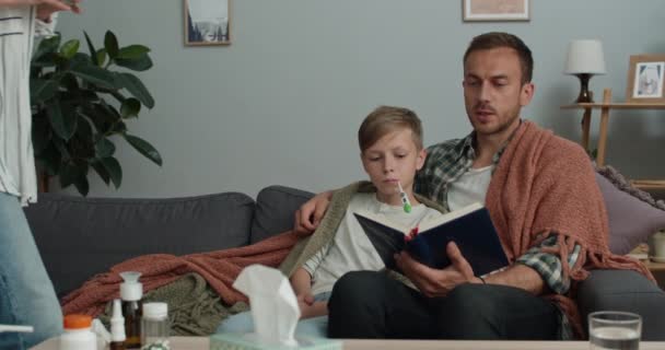 Vader leest boek hardop en zijn zoon meten de temperatuur terwijl ze op de bank zitten.Zorgzame moeder brengen warme thee tijdens het nemen en kijken naar thermometer. Begrip ziekte. — Stockvideo