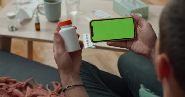 Lvov, Ukrajina - 4. srpna 2020: Pohled přes rameno osoby držící zelenou obrazovku smartphone horizontálně a plastové pilulky láhve zatímco sedí na pohovce doma. Koncept telemedicíny a nemoci. — Stock video