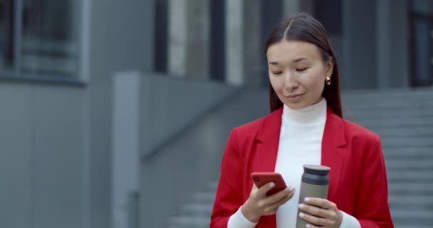 Beskär utsikt över kvinnan med hjälp av smartphone och hålla termo kopp när du står på stadens gata. Attraktiv asiatisk affärskvinna rulla och skriva medan du kommunicerar online. — Stockvideo