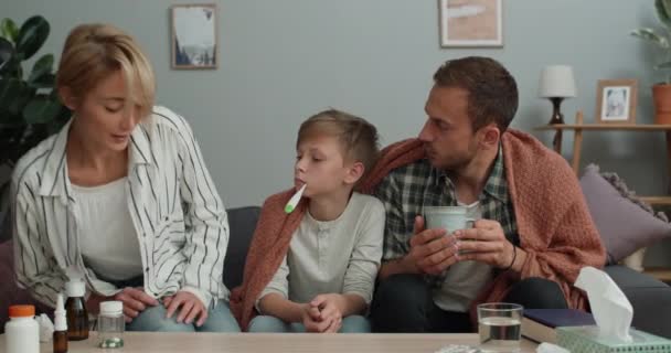 Giovane donna seduta a dare medicine al marito malato e al figlio adolescente. Il padre malato tiene la tazza e il figlio misurano la temperatura mentre si siedono sul divano. Concetto di malattia. — Video Stock