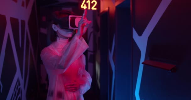 Jovem mulher em roupas da moda vestindo óculos VR em pé no quarto com luzes de néon. Mulher com futurista compõem mãos em movimento enquanto está imersa em realidade virtual. — Vídeo de Stock
