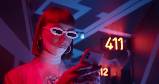 Aproape de o tânără femeie în ochelari de soare la modă tastând mesajul în timp ce stătea pe coridor cu iluminare neon. Femeia cu machiaj futurist folosind smartphone-ul ei . — Videoclip de stoc