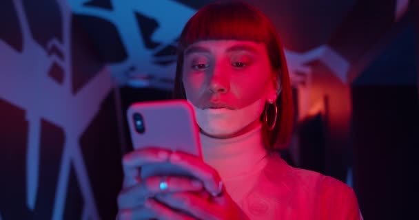 Close up view of beautiful female clubber using smartphone while standing in room with neon lights. Attractive woman with nose ring and futuristic make up chatting in social media. — Stock Video
