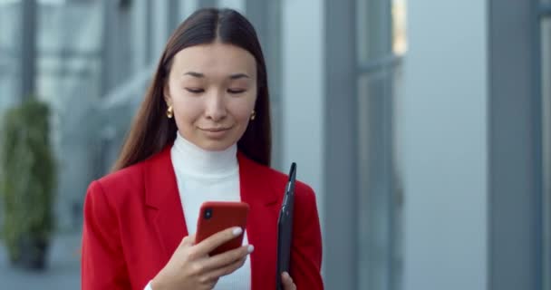 Vedere culturală a femeii CEO navigarea pe internet și zâmbind în timp ce transporta documente dosar de hârtie. Asiatic femeie atractivă folosind smartphone-ul și tastarea în timp ce mersul pe stradă oraș . — Videoclip de stoc