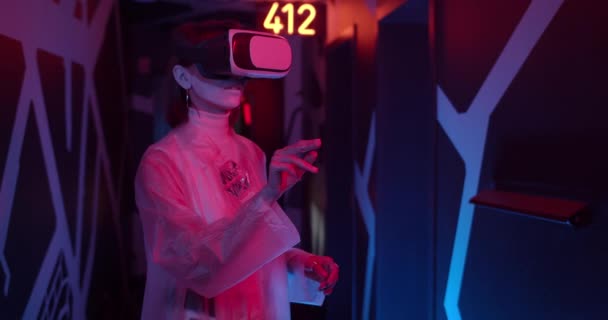 Pemandangan tanaman wanita di headset VR bergerak tangan sambil berdiri di ruangan dengan lampu neon. Perempuan dalam pakaian futuristik mengalami realitas maya. Konsep teknologi modern. — Stok Video