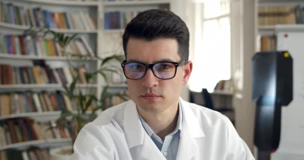 Ritratto di giovane serio che gira la testa e guarda la macchina fotografica mentre si siede nello studio medico. Vista Crop del medico maschile in occhiali e cappotto professionale bianco. Concetto di headshot. — Video Stock