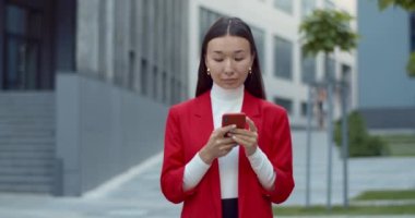 Harika haberler ve gülümsemeyle internete girip şık kıyafetler giyen bir iş kadınının ön görüntüsü. Asyalı kadın şehir caddesinde dururken akıllı telefon ve daktilo kullanıyor..