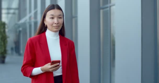 オフィスビルの近くの通りを歩いていると、アジアのビジネスマンの女性が電話の内容をチェックする。スマートフォンやタッチスクリーンを使用して魅力的な若い女性。成功者の概念. — ストック動画