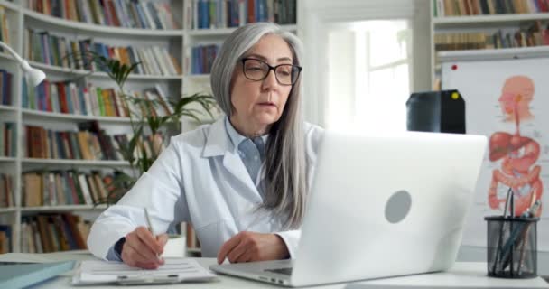 Mujer guapa con gafas y vestido blanco escribiendo y hablando mientras mira la pantalla. Médico anciano que trabaja sentado en la mesa en el consultorio médico. Concepto de telemedicina. — Vídeo de stock