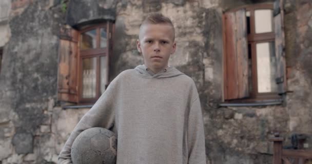 Beskär syn på allvarliga tonåring pojke i luvtröja stående och tittar på kameran. Porträtt av en unge som håller i fotboll när han poserar på gamla stan. Begreppet livsstil och barn. — Stockvideo