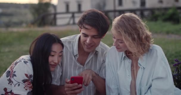 Jonge vrolijke man en twee vrouwen kijken naar het scherm van de telefoon en praten.Glimlachen diverse vrienden met behulp van de smartphone en communiceren tijdens het zitten op gras in het park. Begrip vriendschap. — Stockvideo