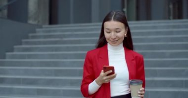 Ofis binasından aşağı inerken elinde kağıt kahve fincanı tutan akıllı telefon kullanan bir kadın görüntüsü. Asyalı iş kadını gülümsüyor, yan tarafa bakıyor ve kameraya bakıyor..