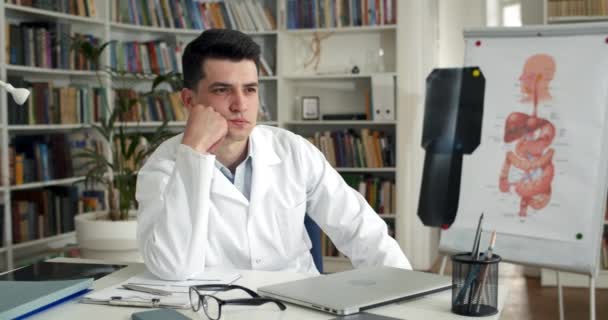 Un tânăr doctor serios se uită la o parte și gândește în timp ce stă la masă cu un laptop pe ea. Bărbat în alb rob profesionist uita atent în timp ce lucrează în cabinetul medical . — Videoclip de stoc