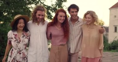 Crop view of millennial friends smiling and embracing together while walking. Group of young people spending time in park. Concept of friendship, unity and leisure.
