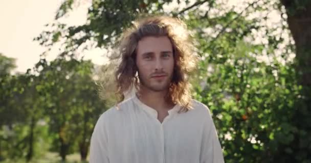 Ritratto di giovane che gira la testa e guarda la macchina fotografica mentre il vento soffia i suoi lunghi capelli biondi. Bello uomo in posa e sorridente mentre in piedi nel parco. Zoom in. — Video Stock